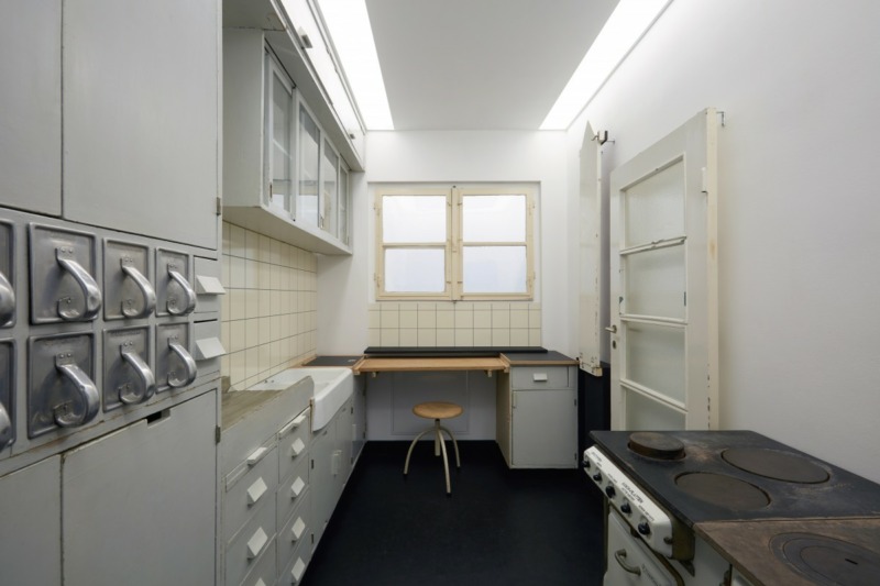 Ausgerichtete Frankfurter Küche in hellen Farben mit Schütten und Schubladen links, Herd und Ofen rechts und Arbeitstisch mit Hocker und Fenster dadrüber. Dazu die Tür. Alle Elemente original, restauriert.