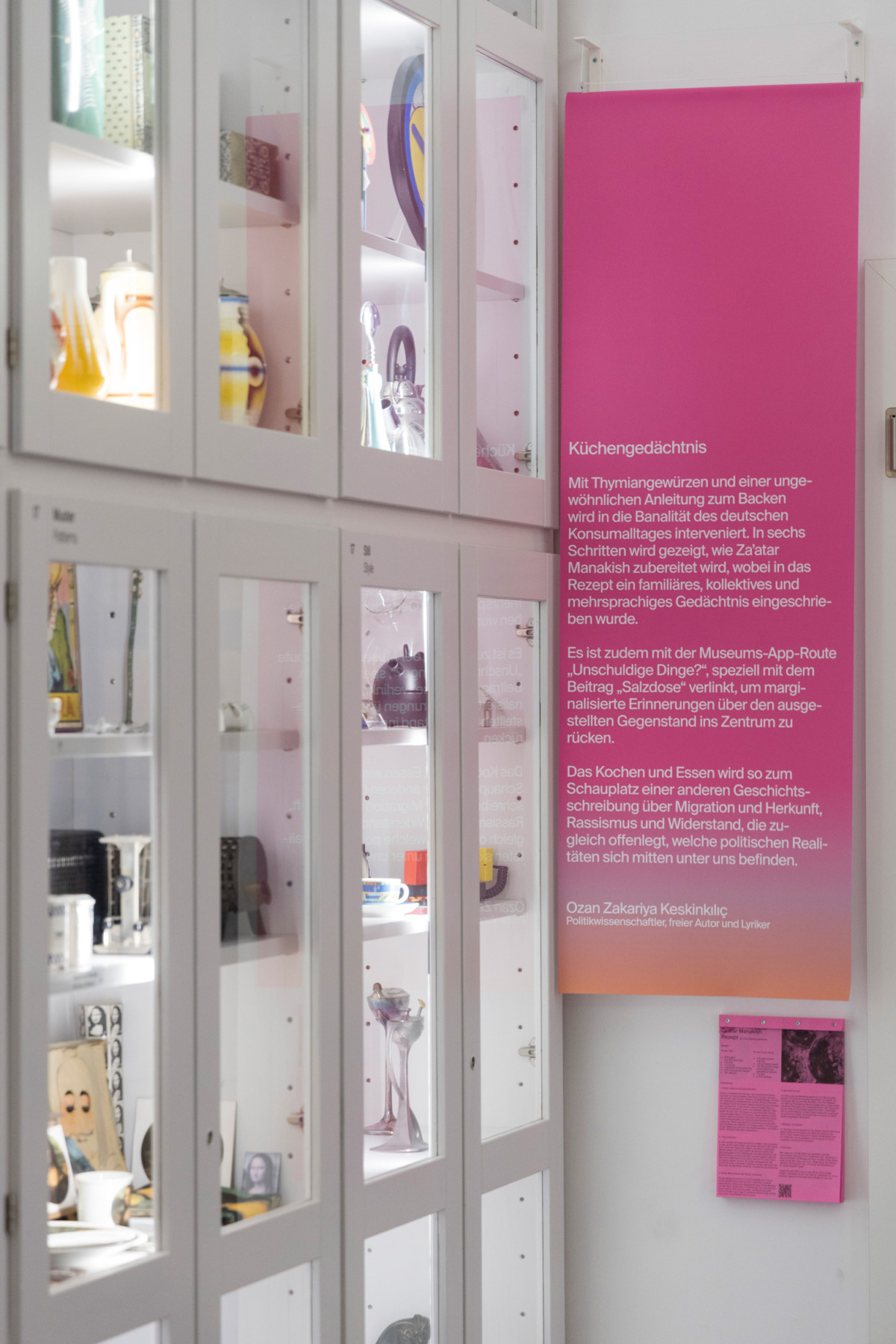 Mit Objekten gefüllte weiße Schränke auf der linken Seite, Rosa gefärbte Fahne auf der rechten Seite.