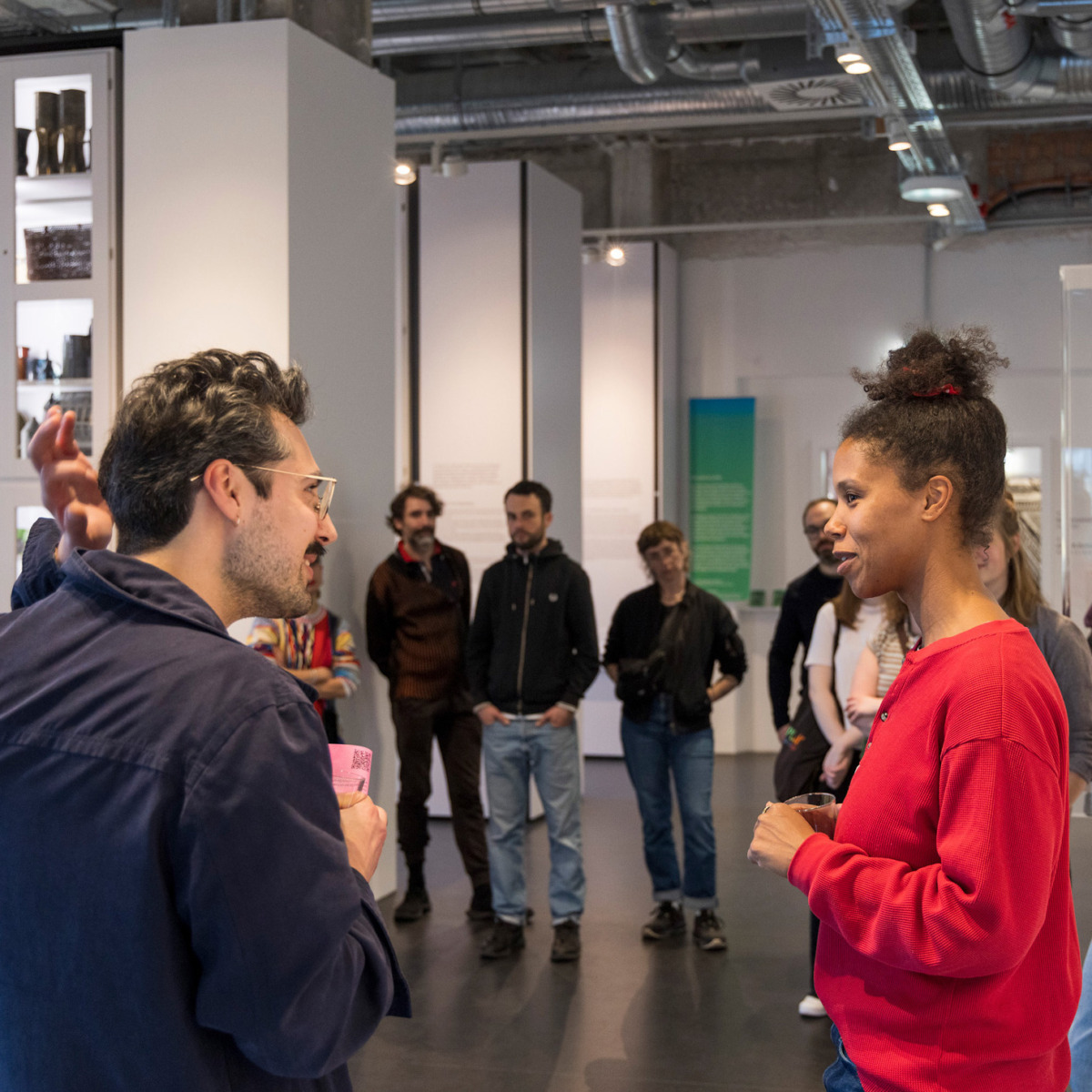 Zwei Personen unterhalten sich in einem Ausstellungsraum. Im Hintergrund weitere Personen, die zuhören.