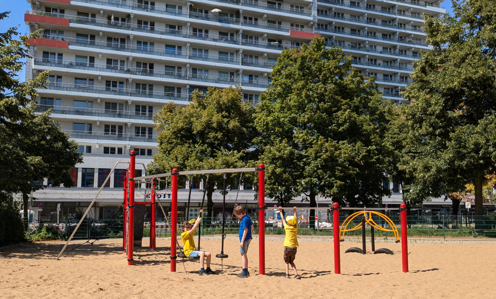 Drei Kinder spielen auf einem Spielplatz vor einem Hochhaus unter scheinender Sonne.