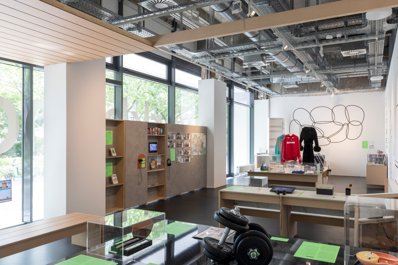 Ausstellungsraum mit verschiedenen Objekten und Möbeln: Kurzhanteln, langen schwarzen vintage-Mantel, Press-on-nails, Tattoo-Sheets in einem Ordner.