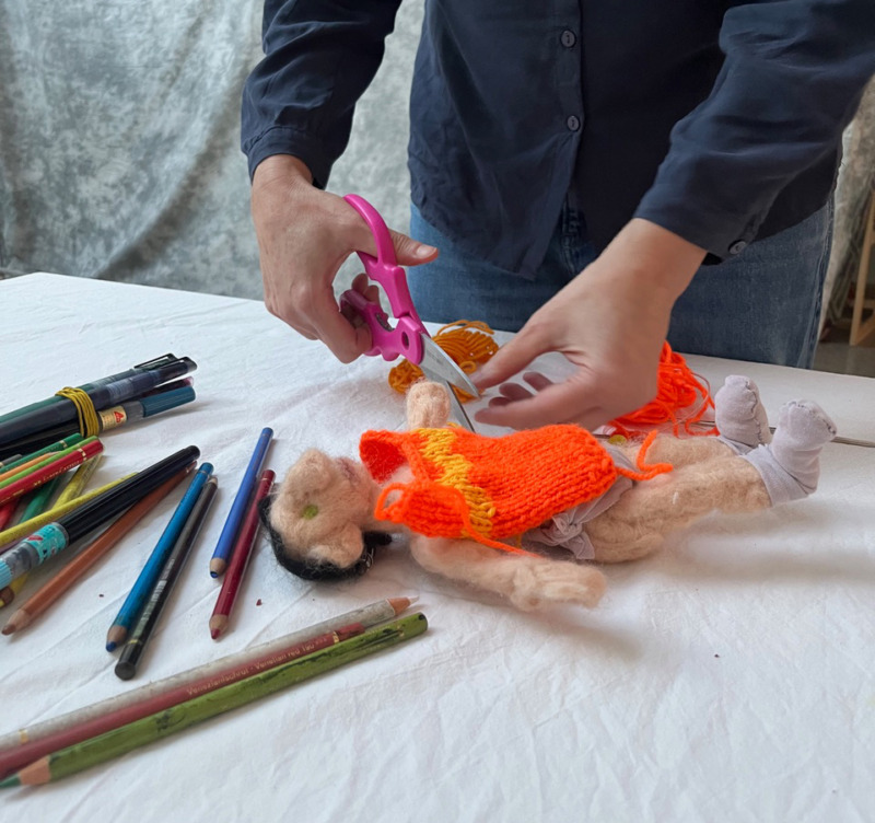 Zwei Hände schneiden Wollfäden, eine Wollpuppe und Buntstifte liegen auf einem tisch
