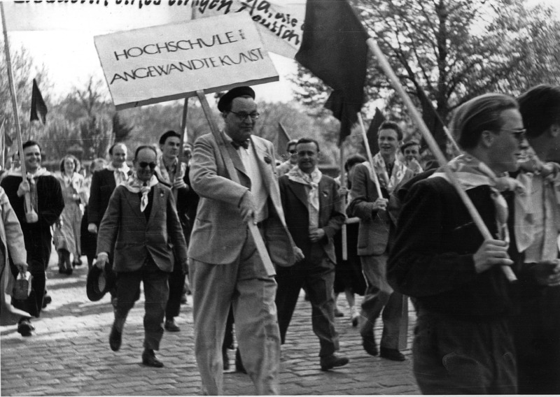 Schwarz-Weiß-Foto von Mart Stam, der mit Baskenmütze an der Spitze einer Demonstration marchiert und ein Schild mit der Aufschrift Hochschule für Angewandte Kunst trägt
