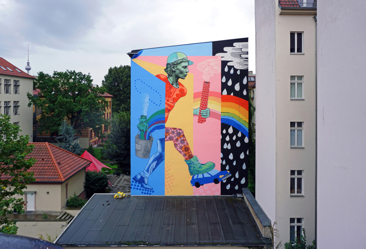 Ein großes, farbenfrohes Wandgemälde an einer Hausfassade zeigt eine rennende Person, die eine rauchende Fabriketage hält, vor einem Hintergrund mit Regenbogen und Regenwetter
