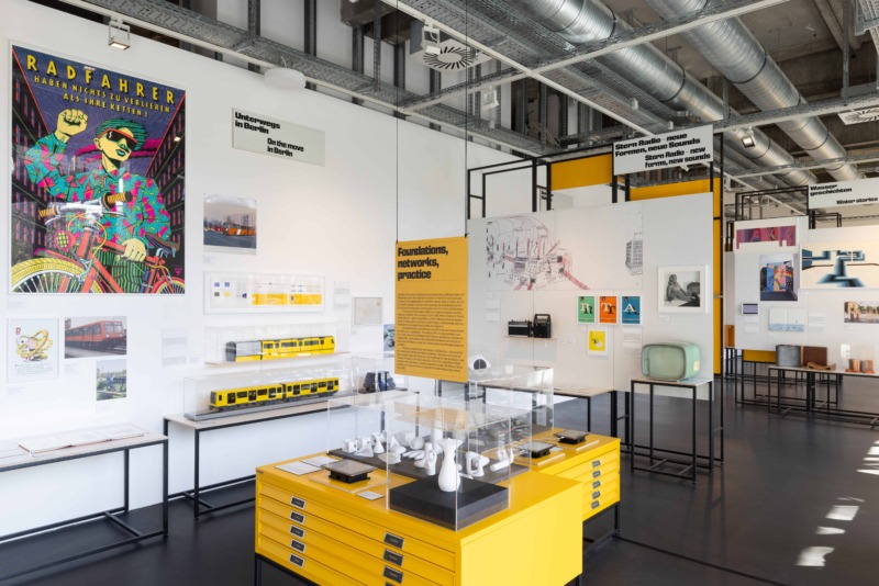 Ausstellungsbereich zum Thema Mobilität in Berlin mit Modellen von U-Bahnen in Vitrinen, einem bunten Plakat 'Radfahrer' und verschiedenen Designobjekten auf gelben Podesten