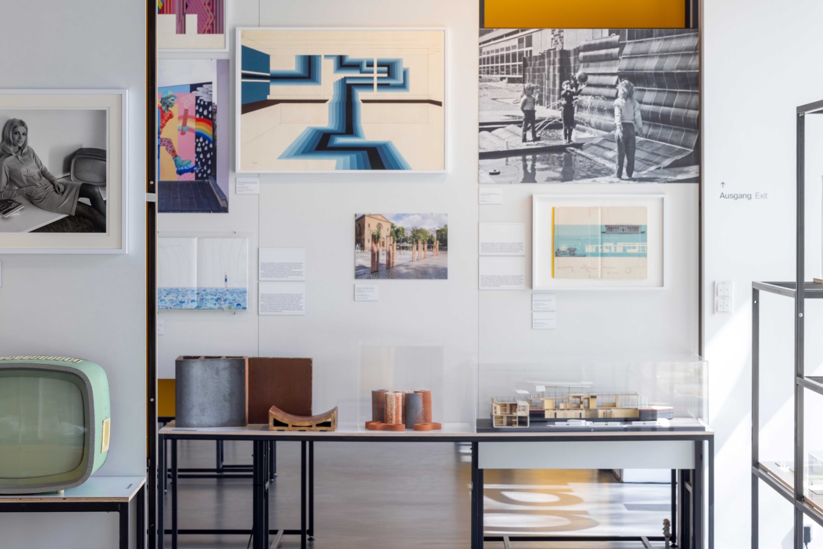 Ausstellungsansicht mit einer weißen Wand voller Architekturzeichnungen und Fotos. Davor ein langer Tisch mit Architekturmodellen in Vitrinen sowie Materialproben. Links ein hellgrüner Retro-Fernseher.