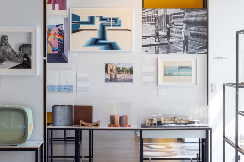 Ausstellungsansicht mit einer weißen Wand voller Architekturzeichnungen und Fotos. Davor ein langer Tisch mit Architekturmodellen in Vitrinen sowie Materialproben. Links ein hellgrüner Retro-Fernseher.
