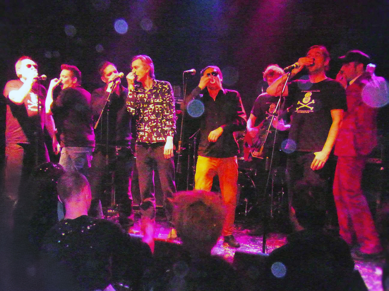 Eine mehrköpfige Band singt und performt live auf einer Clubbühne unter buntem Scheinwerferlicht