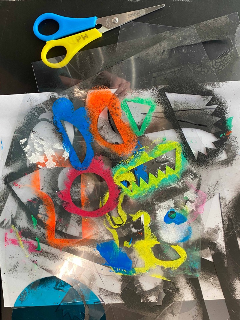 Verschiedene bunte Schablonen aus transparentem Kunststoff liegen übereinander auf einem mit Farbe besprühten Papier