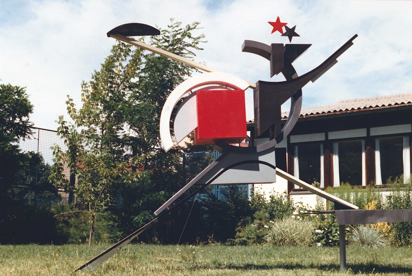 Eine avantgardistische, konstruktivistische Skulptur mit abstrakten geometrischen Formen wie einem roten Quadrat und zwei Sternen an der Spitze steht auf einer Rasenfläche im Freien.